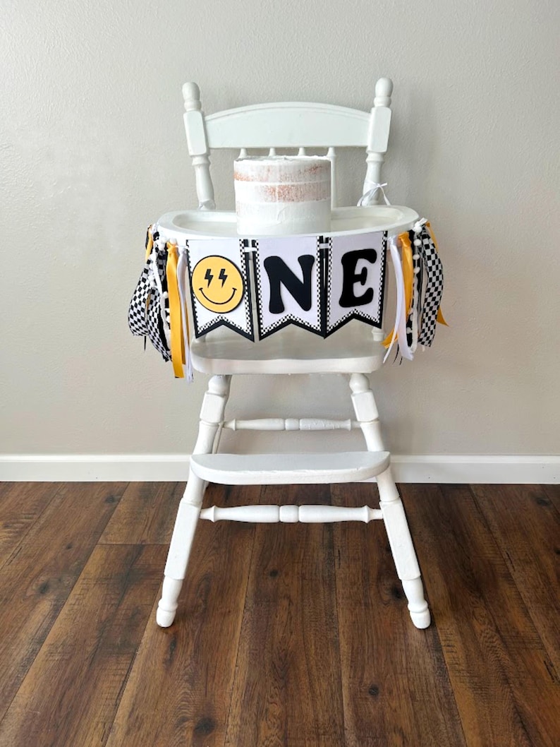 May include: A white high chair decorated for a first birthday. The chair has a banner that says "ONE" with a smiley face and lightning bolt graphic. The banner is black and white with yellow accents. A small cake sits on the tray.