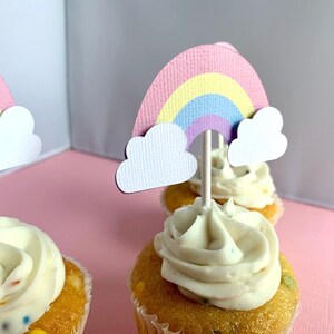 May include: Three cupcakes with white frosting and rainbow sprinkles topped with paper rainbow and cloud cupcake toppers.