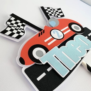 May include: Red race car cake topper with a checkered flag, the number 2, and the name Theo in blue letters.