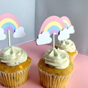 May include: Three cupcakes with white frosting and rainbow sprinkles topped with rainbow and cloud cupcake toppers.