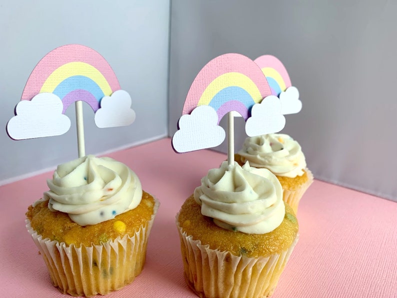 May include: Three cupcakes with white frosting and rainbow sprinkles topped with rainbow and cloud cupcake toppers.