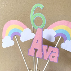 May include: A cake topper set featuring a pink "Ava" name, a green number "6", and two rainbow and cloud designs. The topper is ideal for a birthday party.