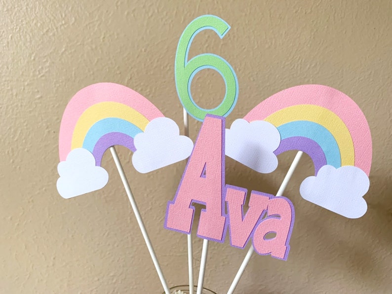May include: A cake topper set featuring a pink "Ava" name, a green number "6", and two rainbow and cloud designs. The topper is ideal for a birthday party.