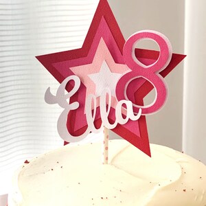May include: A pink and red layered star cake topper with the name "Ella" and the number "8" on it. The topper is on a white frosted cake.