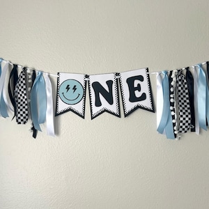 May include: A blue and black "ONE" banner with a smiley face with lightning bolts for eyes. The banner is decorated with blue, white, and black ribbons.