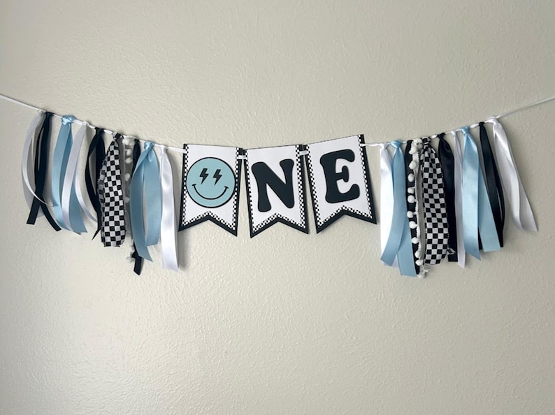 May include: A blue and black "ONE" banner with a smiley face with lightning bolts for eyes. The banner is decorated with blue, white, and black ribbons.