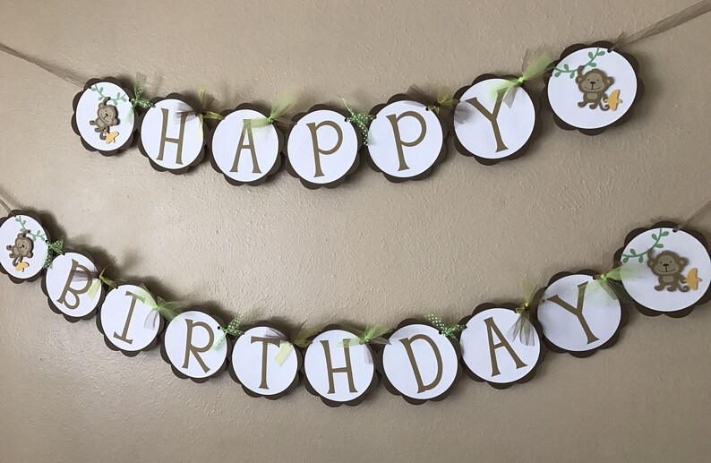 May include: A brown and white banner with the words "Happy Birthday" decorated with green leaves and small monkeys. The banner is strung with twine and has brown scalloped edges.