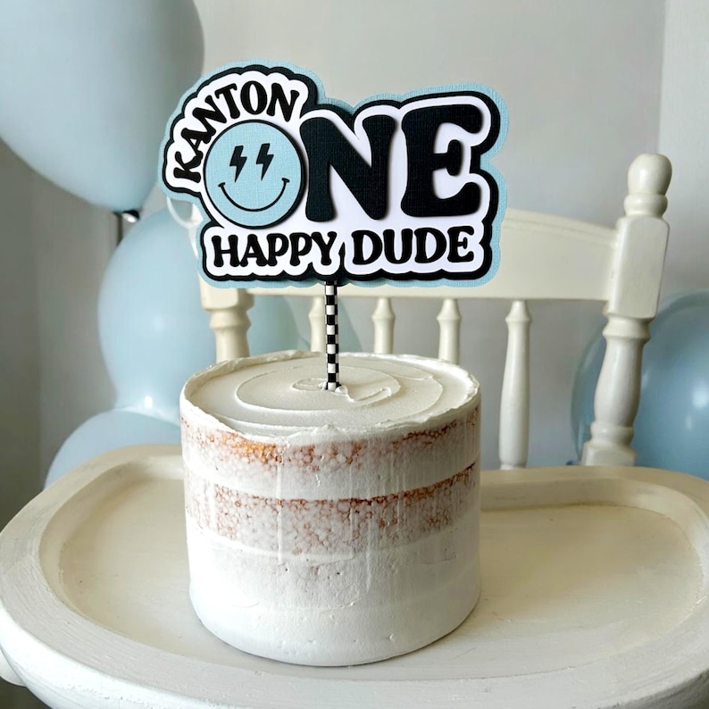 May include: A white cake with white frosting and a blue and black cake topper that says "Kanton One Happy Dude" with a smiley face.