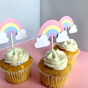 May include: Three cupcakes with white frosting and rainbow sprinkles topped with pastel rainbow cupcake toppers. The toppers are made of paper and have a cloud shape at the bottom.