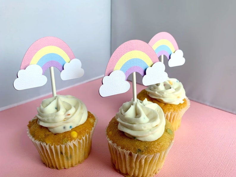 May include: Three cupcakes with white frosting and rainbow sprinkles topped with pastel rainbow cupcake toppers. The toppers are made of paper and have a cloud shape at the bottom.