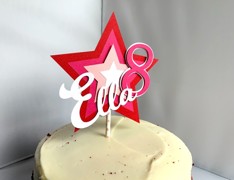 May include: A red, pink and white star-shaped cake topper with the name "Ella" and the number "8" in pink. The topper is on a white frosted cake.