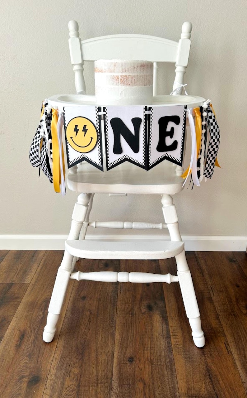 White, black and yellow cardstock banner with yellow and black smile face and black N and E with ribbons hanging on sides attached to a white high chair.