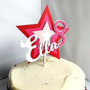 May include: A red, pink and white star-shaped cake topper with the name "Ella" and the number "8" on it. The topper is on a white frosted cake.