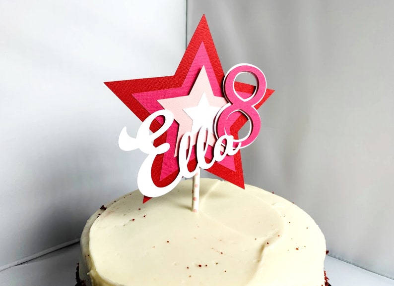 May include: A red, pink and white star-shaped cake topper with the name "Ella" and the number "8" on it. The topper is on a white frosted cake.