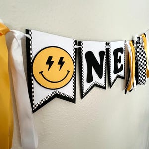 May include: A banner with the word "ONE" in black letters. The first banner has a yellow smiley face with lightning bolts. The banners are white with a black and white checkered border. Yellow and white ribbons hang from the banner.