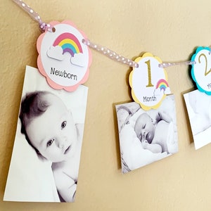 May include: A rainbow-themed baby milestone banner with pink, yellow, and blue circles. Each circle features a number and a rainbow with clouds. The banner is decorated with white polka dot ribbon. Photos of a baby are attached to the banner.