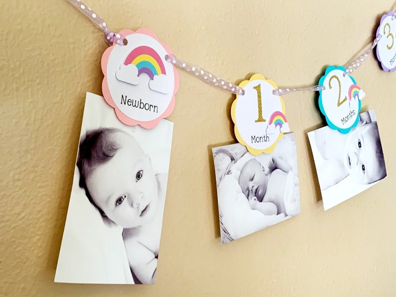 May include: A rainbow-themed baby milestone banner with pink, yellow, and blue circles. Each circle features a number and a rainbow with clouds. The banner is decorated with white polka dot ribbon. Photos of a baby are attached to the banner.
