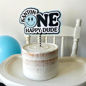 May include: A blue and white cake topper with the text "Kanton One Happy Dude" and a smiley face with lightning bolts. The topper is on a white cake with white frosting.