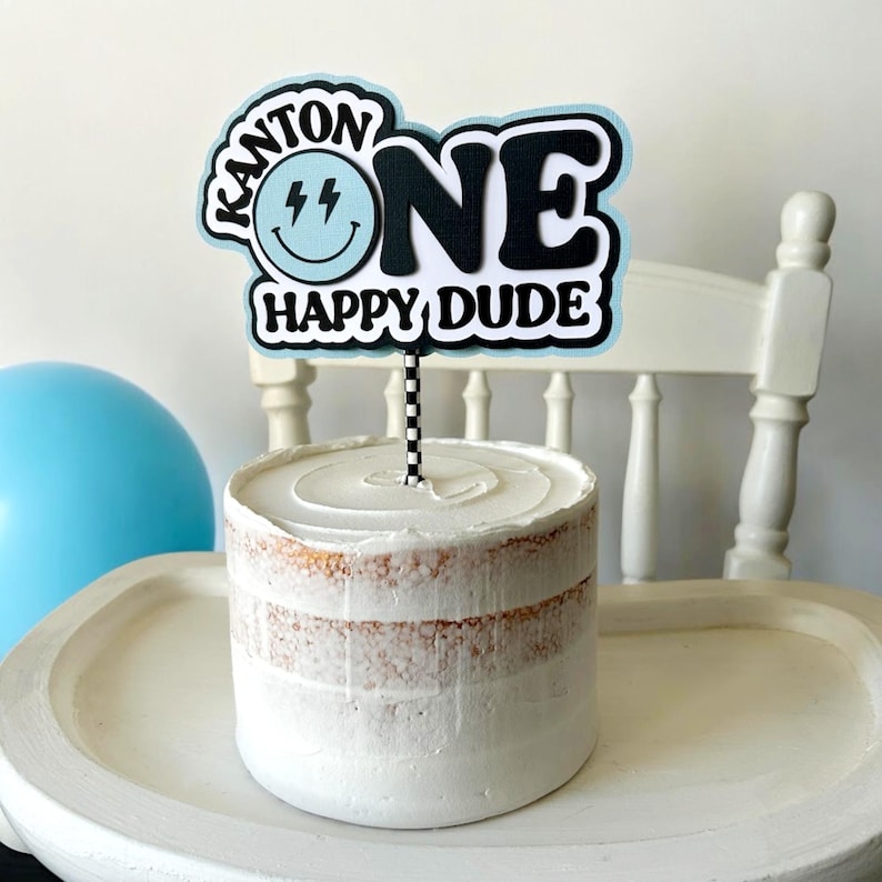 May include: A blue and white cake topper with the text "Kanton One Happy Dude" and a smiley face with lightning bolts. The topper is on a white cake with white frosting.
