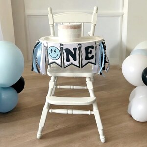 May include: A white high chair with a black and white banner that says "ONE" with a blue and black smiley face on the left side. The banner has black and white striped ribbon streamers hanging from it. There is a white cake on the tray of the high chair. There are blue and white balloons in the background.