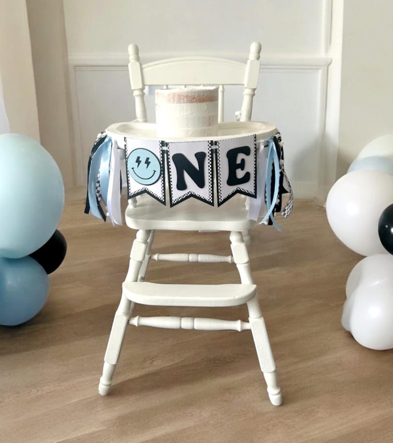 May include: A white high chair with a black and white banner that says "ONE" with a blue and black smiley face on the left side. The banner has black and white striped ribbon streamers hanging from it. There is a white cake on the tray of the high chair. There are blue and white balloons in the background.