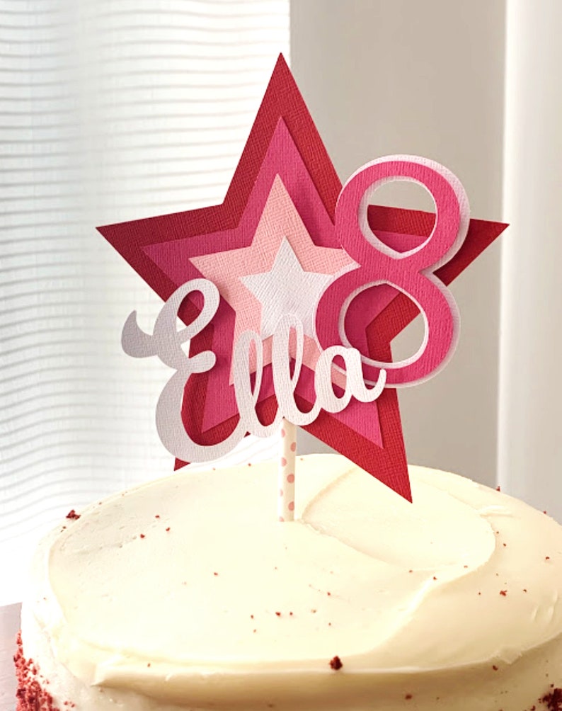 May include: A pink and white layered star cake topper with the name "Ella" and the number "8" on it. The topper is on a white cake with red frosting.