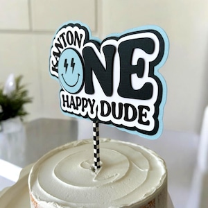 May include: A blue and black cake topper with the word "ONE" in large black letters and the text "Happy Dude" below. The topper also features a blue smiley face with lightning bolts.