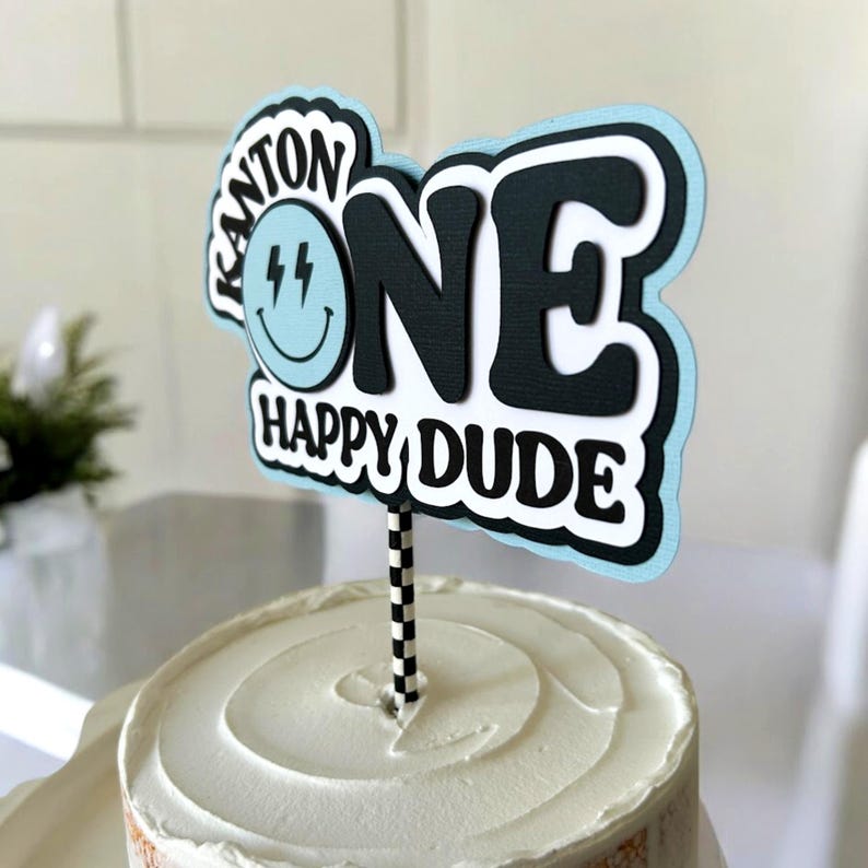 May include: A blue and black cake topper with the word "ONE" in large black letters and the text "Happy Dude" below. The topper also features a blue smiley face with lightning bolts.