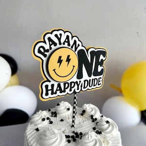 May include: A cake topper with the text "RAYAN ONE HAPPY DUDE" in black and white, with a yellow smiley face and lightning bolts. The topper is on a black and white striped stick, placed on a white frosted cake with chocolate chips. Balloons are in the background.