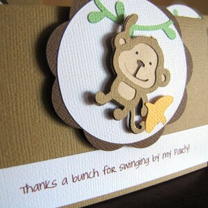 May include: A brown card with a white circle in the center. The circle has a brown border and a scalloped edge. Inside the circle is a cartoon monkey swinging from a vine. The monkey is brown with a yellow banana. The card has the text "Thanks a bunch for swinging by my party!" printed on it.