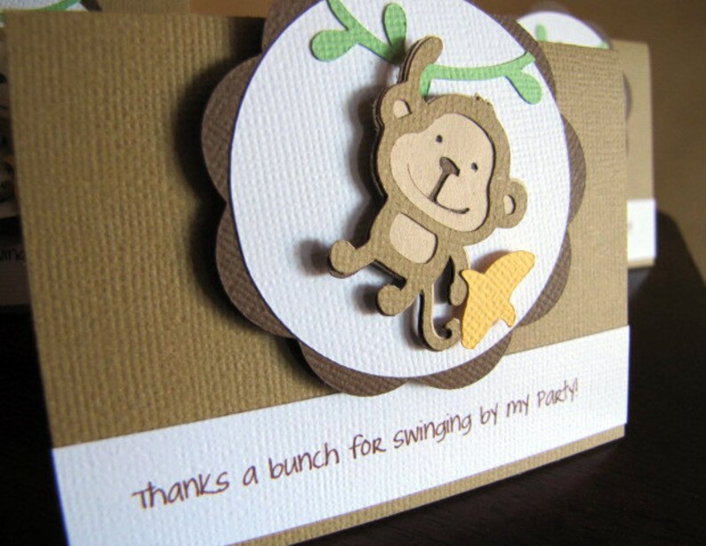May include: A brown card with a white circle in the center. The circle has a brown border and a scalloped edge. Inside the circle is a cartoon monkey swinging from a vine. The monkey is brown with a yellow banana. The card has the text "Thanks a bunch for swinging by my party!" printed on it.