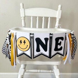 May include: White high chair decorated with a banner that says "ONE". The banner has a yellow smiley face with lightning bolts, black and white checkered flags, and yellow and white ribbons. The high chair is made of wood.