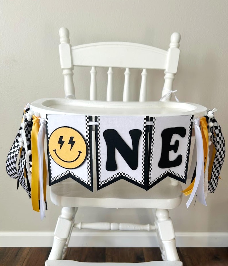 May include: White high chair decorated with a banner that says "ONE". The banner has a yellow smiley face with lightning bolts, black and white checkered flags, and yellow and white ribbons. The high chair is made of wood.
