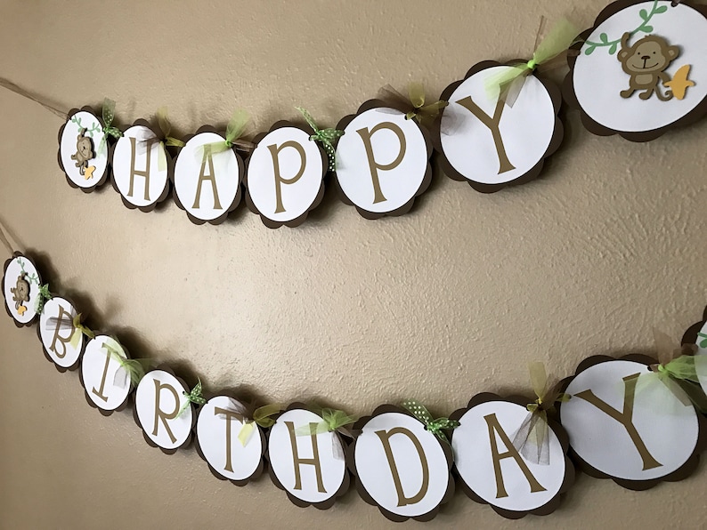 Może przedstawiać: Brązowo-biały baner z napisem "Happy Birthday" złotymi literami. Baner jest ozdobiony zieloną i ż&oacute;łtą wstążką oraz małymi figurkami małp.