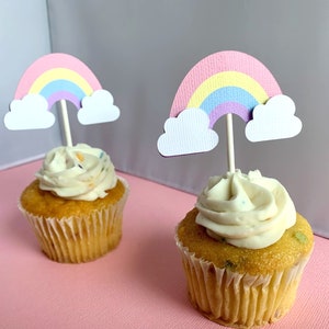 May include: Two cupcakes with white frosting and rainbow sprinkles topped with rainbow and cloud cupcake toppers.