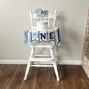 May include: A white high chair with a blue and black banner that says "ONE" and a cake topper that says "KANTOR ONE HAPPY DUDE".