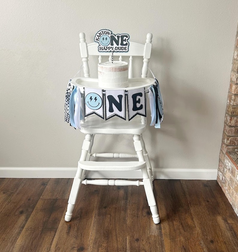 May include: A white high chair with a blue and black banner that says "ONE" and a cake topper that says "KANTOR ONE HAPPY DUDE".