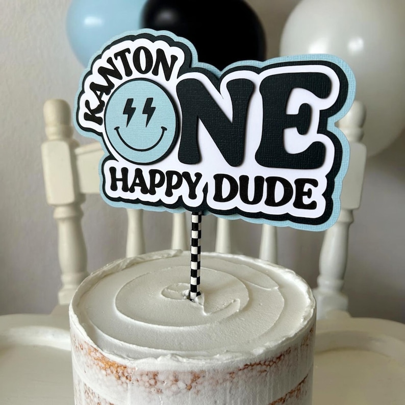 May include: A blue and black cake topper with the text "Kanton One Happy Dude" and a smiley face with lightning bolts for eyes.