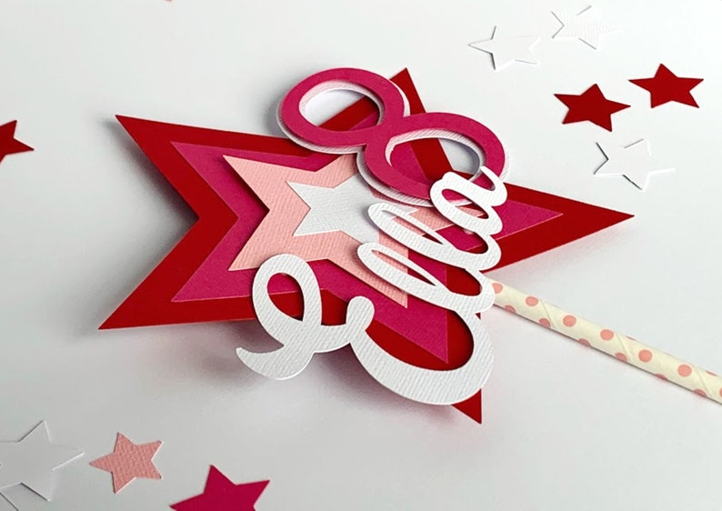 May include: A pink and red layered star with the name "Ella" and the number "8" cut out of white paper. The star is attached to a pink and white striped straw.