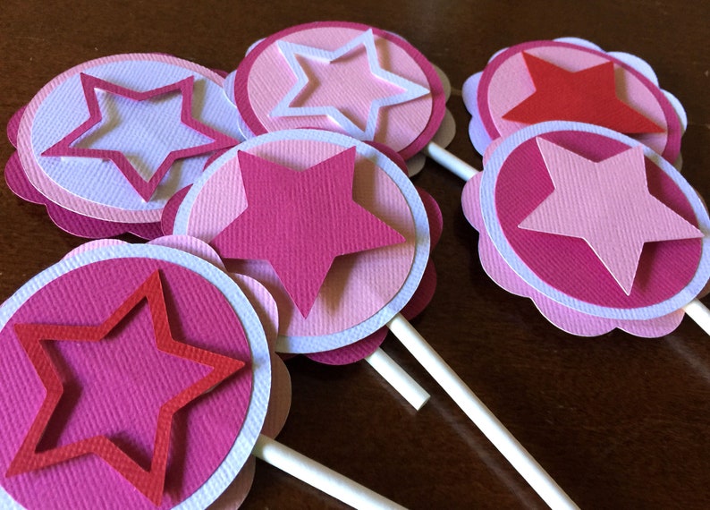 May include: Six pink and white paper cupcake toppers with a star design. The stars are layered with different shades of pink and white.