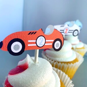 May include: Three cupcakes decorated with red, white, and blue racing car toppers. The toppers are made of paper and feature the numbers "2" and "1".