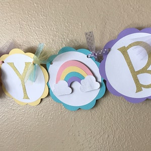 May include: Happy Birthday banner with scalloped circles in pink, yellow, turquoise and purple. Each circle has a white background with gold glitter letters and a rainbow with clouds on one circle.