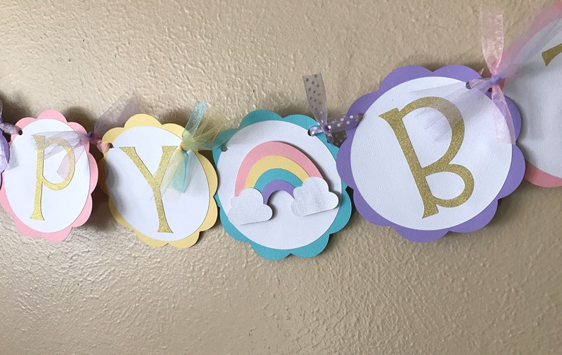 May include: Happy Birthday banner with scalloped circles in pink, yellow, turquoise and purple. Each circle has a white background with gold glitter letters and a rainbow with clouds on one circle.