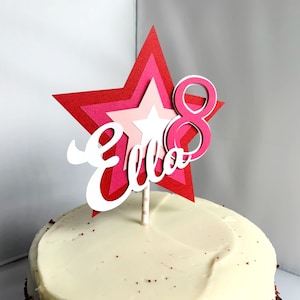 May include: A pink and red layered star cake topper with the name "Ella" and the number "8" on it. The topper is on top of a white frosted cake.