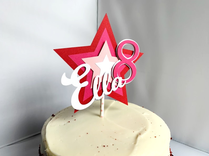 May include: A pink and red layered star cake topper with the name "Ella" and the number "8" on it. The topper is on top of a white frosted cake.