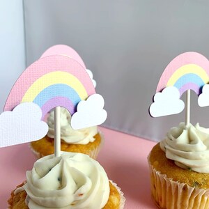 May include: Three cupcakes topped with rainbow and cloud decorations. The cupcakes are frosted with white icing and have sprinkles. The rainbow toppers are made of paper and feature a pastel colour scheme.