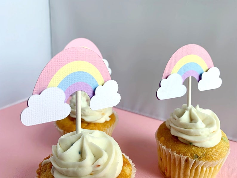 May include: Three cupcakes topped with rainbow and cloud decorations. The cupcakes are frosted with white icing and have sprinkles. The rainbow toppers are made of paper and feature a pastel colour scheme.