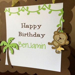 May include: A brown and green birthday banner with a white background. The banner has a green vine border with leaves and a monkey hanging from it. The banner says "Happy Birthday Benjamin".