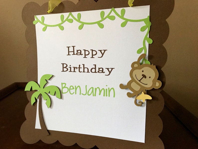 May include: A brown and green birthday banner with a white background. The banner has a green vine border with leaves and a monkey hanging from it. The banner says "Happy Birthday Benjamin".