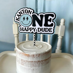 May include: A blue and white cake topper with the text "Kanton One Happy Dude" and a lightning bolt smiley face. The topper is on a white cake.